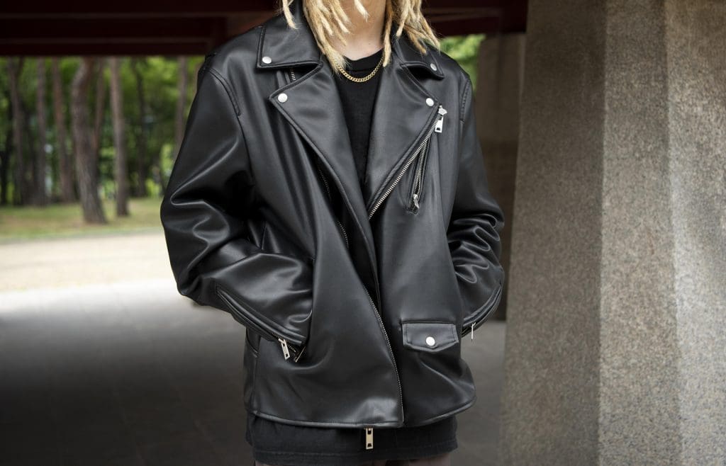 Person wearing a black leather jacket and black shirt stands outdoors with hands in jacket pockets; head is partially out of frame.