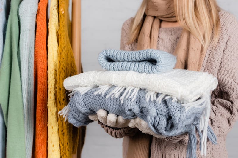A person wearing a scarf and gloves holds a stack of folded knit blankets and sweaters, with more colorful knitwear hanging in the background.